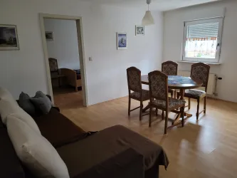 Ein gemütlicher Wohn- und Essbereich mit einem runden Tisch und vier Stühlen. Die Wände sind hell, und es gibt ein großes Fenster, das natürliches Licht hineinlässt.