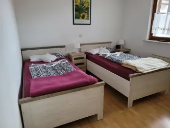 Ein einfaches Schlafzimmer mit zwei Einzelbetten und Holzmöbeln. Die Wände sind hell gestrichen und ein Bild hängt darüber.