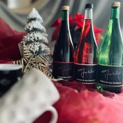 Drei Flaschen Glühwein in verschiedenen Farben stehen auf einem festlichen Untergrund mit roten Blüten. Im Hintergrund sind Weihnachtsdekorationen und eine kleine Tanne zu sehen.