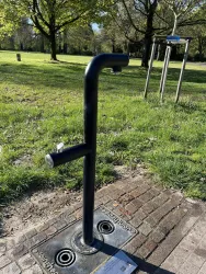 Ein Trinkbrunnen in einem Park steht auf einem gepflasterten Boden. Im Hintergrund sind Bäume und eine Wiese zu sehen.