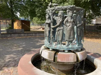 Ein schöner Brunnen mit einer Skulptur von mehreren Figuren, die Traditionen darstellen. Umgeben von Bäumen und einem gepflasterten Platz.