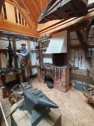 Eine historische Schmiede mit einem Schmied und vielen Werkzeugen. Der Raum ist rustikal und gut eingerichtet, mit einem Herd und einem Amboss.