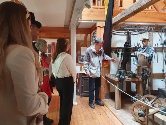 Eine Gruppe von Menschen steht in einem Museum und hört einem Guide zu, der auf eine alte Maschine zeigt. Im Hintergrund sind historische Objekte und ein deutscher Fahnenmast zu sehen.
