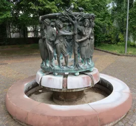 Ein kunstvoller Brunnen mit Figuren, die um einen Baum gruppiert sind. Die Statue zeigt Menschen in traditoneller Kleidung, umgeben von einer grünen Umgebung.