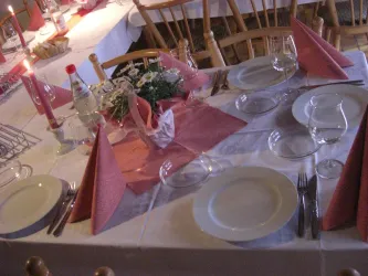 Ein elegant gedeckter Tisch mit weißen Tellern, Gläsern und rosanen Servietten. Auf dem Tisch stehen Kerzen und eine kleine Blumendekoration.