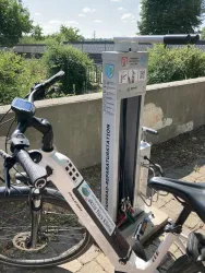 Eine Fahrradreparaturstation steht neben einem Fahrrad. Im Hintergrund sind Bäume und ein blauer Himmel zu sehen.
