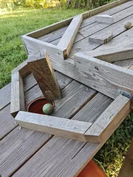 Ein Holzspielgerät mit einer mehrfachen Struktur und bunten Kugeln liegt auf einem Tisch. Der Hintergrund zeigt grünes Gras und Natur.