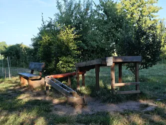 Eine ruhige Gartenlandschaft mit einer Bank und einem einfachen Spielbereich. Umgeben von Bäumen und grünem Gras.