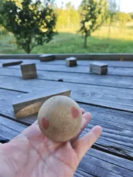 Eine Hand hält eine hölzerne Kugel mit zwei roten Herzen. Im Hintergrund sind Bäume und ein Holztisch zu sehen.