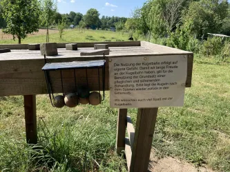 Eine Kugbahn aus Holz steht in einer grünen Umgebung. Schilder informieren über die Nutzung und das Spielen mit den Kugeln.