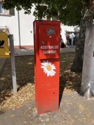 Eine rote Säule mit der Aufschrift "KOSTENLOS TANKEN!" und einer weißen Blume. Sie steht auf einem Gehweg im Freien, umgeben von Bäumen.