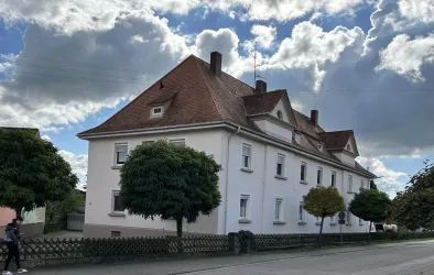 Ein großes, weißes Gebäude mit einem roten Dach und großen Fenstern. Mehrere Bäume stehen im Vordergrund vor einem bewölkten Himmel.