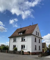 Ein weißes Haus mit einem braunen Dach und mehreren Fenstern. Der Himmel ist blau und mit einigen Wolken bedeckt.