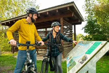 Ein Paar mit Fahrrädern steht an einer Informationstafel in der Natur. Sie schauen sich die Informationen an und diskutieren wahrscheinlich über die Umgebung.