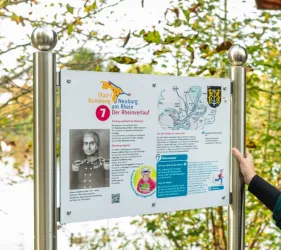 Ein Wegweiser für den Radweg in Neuburg am Rhein zeigt Informationen über den Rheinuferlauf. Die Tafel enthält eine Karte und historische Informationen sowie ein Bild einer Person.