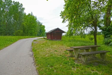Eine ruhige Landschaft mit einem Weg und einer roten Hütte. Neben dem Weg stehen Holzbank und Tisch unter einem Baum.