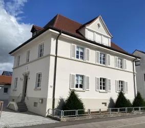Ein modernes, zweistöckiges Haus mit weißen Wänden und roten Dachziegeln. Vor dem Haus stehen einige kleine immergrüne Bäume.