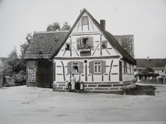 Ein traditionelles Fachwerkhaus mit einem offenen Fenster und einem großen Tor. Vor dem Haus stehen fünf Personen.