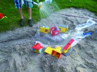 Jemand gießt Wasser mit einer Gießkanne über einen im Sandkaten gebauten Graben, an dessen Ufern Häuser aus Lego stehen