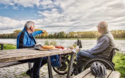 Zwei Männer, einer davon im Rollstuhl, sitzen an einem barrierefreien Rastplatz direkt am Rheinufer