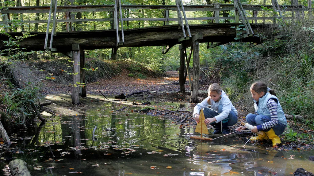 Zwei Mädchen spielen mit einem kleinen Segelboot an einem flachen Bach im Wald