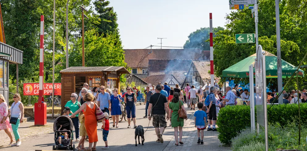 Viele Festbesucher spazieren bei herrlichem Wetter über eine für den Verkehr gesperrte Straße. Im Hintergrund sind Buden uns große Sonnenschirme zu erkennen.