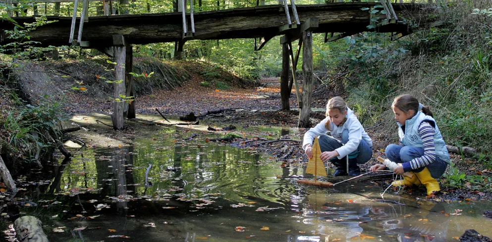 Zwei Mädchen spielen mit einem kleinen Segelboot an einem flachen Bach im Wald