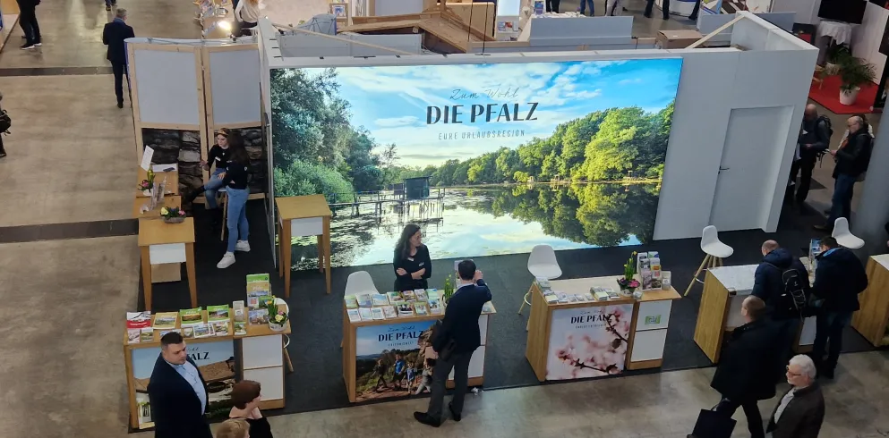 Messestand auf der CMT, im Hintergrund ein hell erleuchtetes Bild der Rheinauen, vornedran 5 Counter mit Angestellten und einigen Gästen, die sich unterhalten.