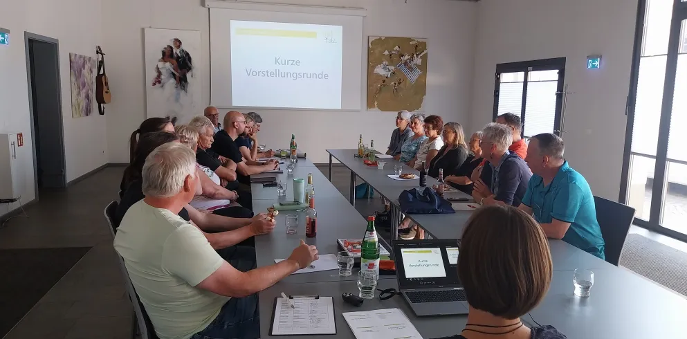 Eine Gruppe von ca. 20 Personen sitzt sich an Tischen gegenüber und schaut nach vorne auf eine Präsentation