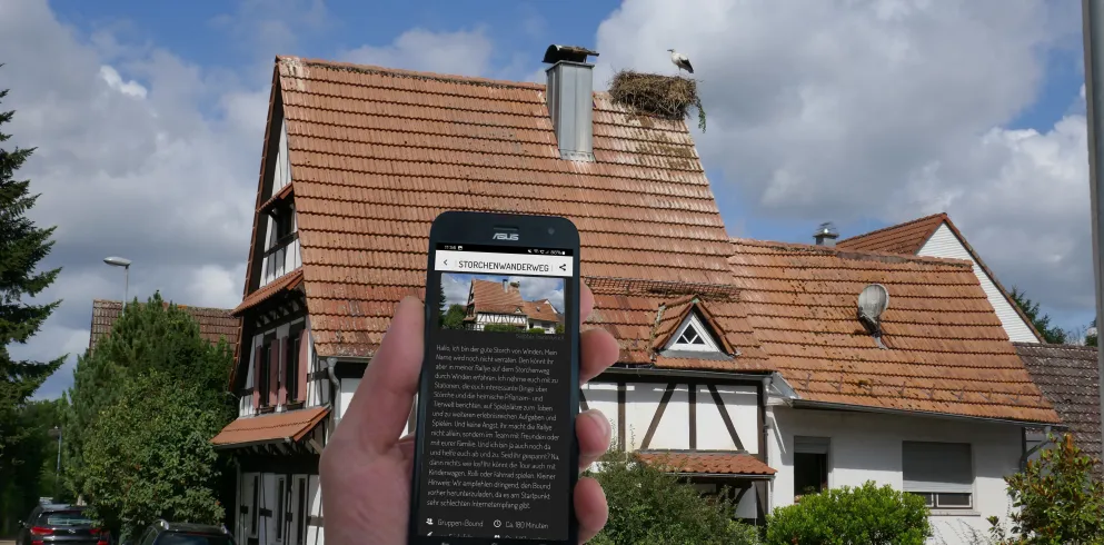 Eine Hand mit Smartphone, auf welchem das Storchenquiz geöffnet ist, vor dem Hintergrund eines Fachwerkhauses mit Storchenhorst.