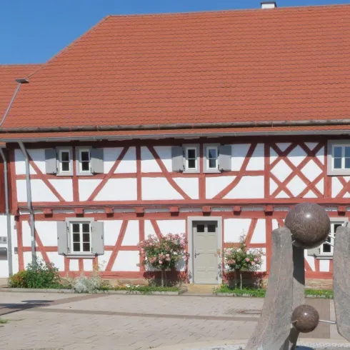 Ein altes Fachwerkhaus mit rotem Dach und weißen Wänden steht in einem ruhigen Platz. Neben dem Haus befindet sich eine moderne Skulptur und blühende Pflanzen.