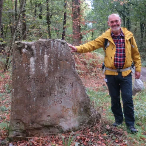 Ein Mann in gelber Jacke steht neben einem großen Stein in einem Wald. Der Stein trägt eine Inschrift und das Umfeld ist herbstlich mit bunten Blättern.