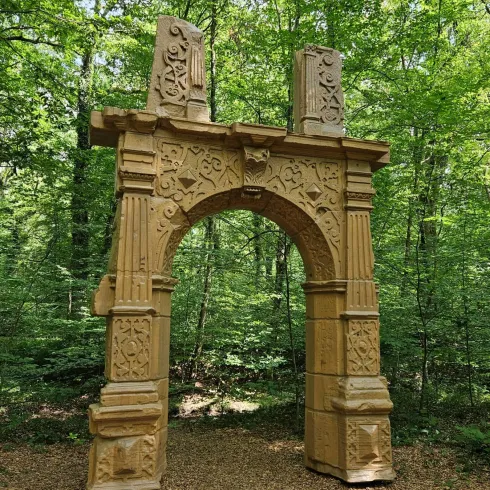 Ein kunstvoll gestalteter Bogen steht im Wald zwischen Bäumen. Der Bogen hat zahlreiche Verzierungen und ist aus hellem Material gefertigt.