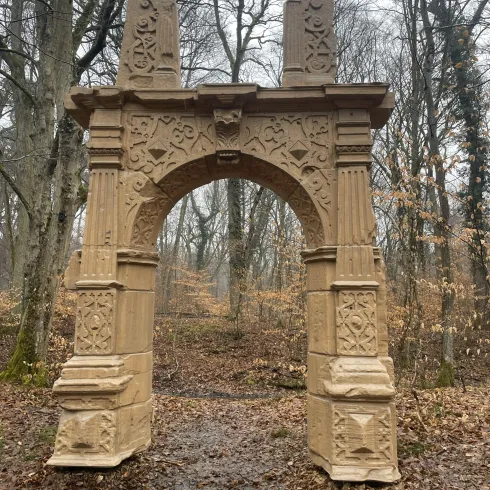 Ein dekorativer Bogen aus Stein steht in einem Wald. Umgeben von Bäumen und herbstlichem Laub.