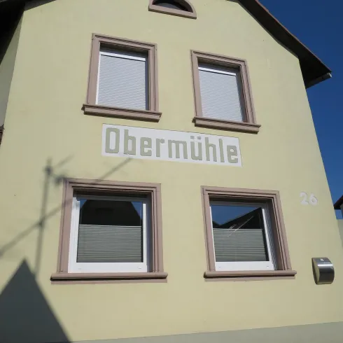 Ein gelbes Gebäude mit dem Schriftzug "Obermühle". Es hat drei Fenster und einen blauen Himmel im Hintergrund.