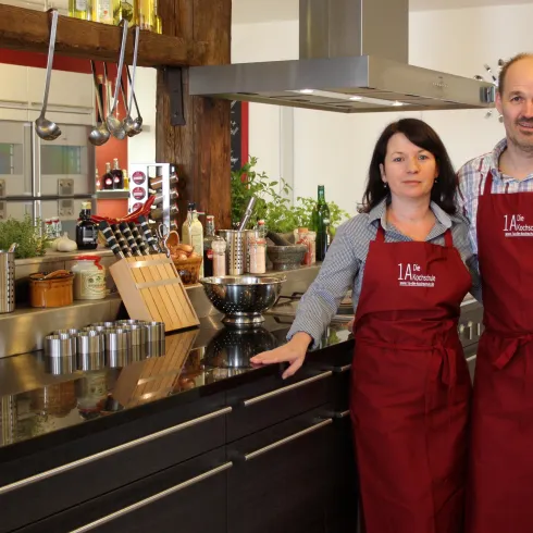 Ein modernes Küchensetting mit zwei Personen in Schürzen. Im Hintergrund sieht man frische Kräuter und Kochutensilien.