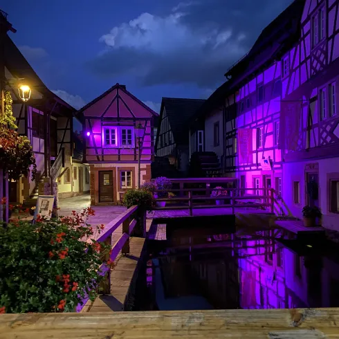 Eine malerische Altstadt mit Fachwerkhäusern und einem kleinen Wasserlauf. Die Szene wird von lila Beleuchtung und einem stimmungsvollen Himmel bei Nacht geprägt.