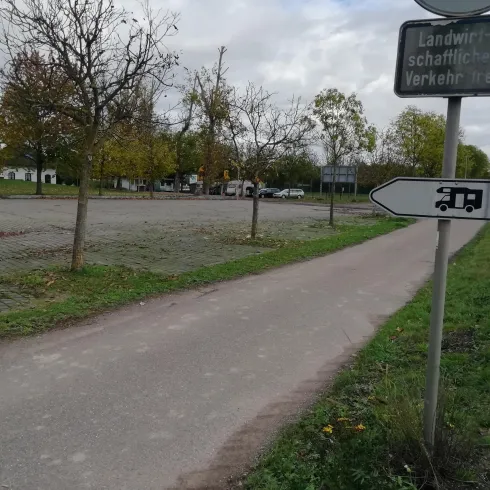 Ein Parkplatz mit einigen Bäumen und einer Fahrbahn. Ein Wegweiser zeigt in Richtung eines Wohnmobils.