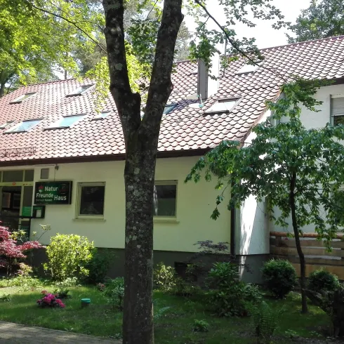 Ein modernes Gebäude umgeben von Bäumen und blühenden Sträuchern. Es hat ein rotes Satteldach und eine Zugangsrampe.