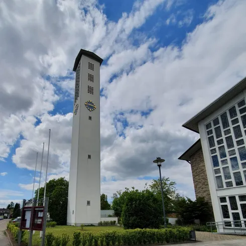 Ein hoher, moderner Turm steht neben einem Gebäude unter einem klaren, blauen Himmel mit weißen Wolken. Üppiges Grün umgibt die Umgebung.