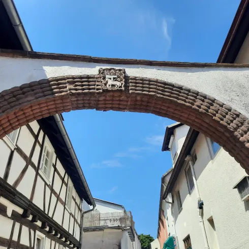 Ein alter, bogenförmiger Eingang aus Ziegelsteinen, der zwischen zwei Häusern hindurchführt. Darüber ist ein Wappen in das Mauerwerk eingearbeitet, und der Himmel ist klar und blau.