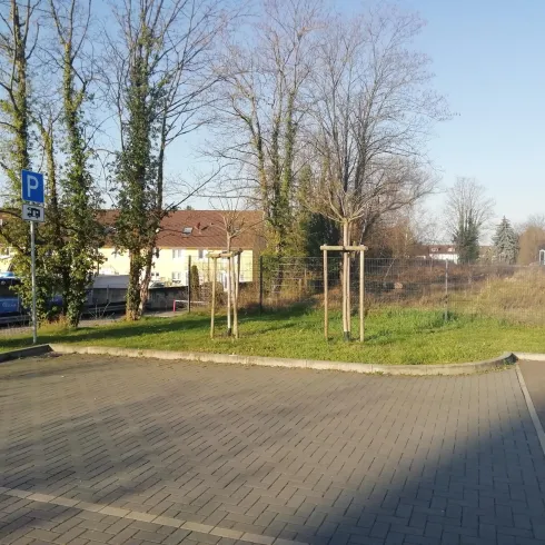 Ein Parkplatz mit gepflastertem Boden und Bäumen im Hintergrund. Die Umgebung ist freundlich und hell, mit klaren Himmel und Wohngebäuden in der Nähe.