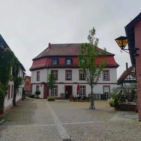 Ein schöner Platz mit einem traditionellen Haus und grünen Bäumen. Die Atmosphäre ist ruhig und einladend.