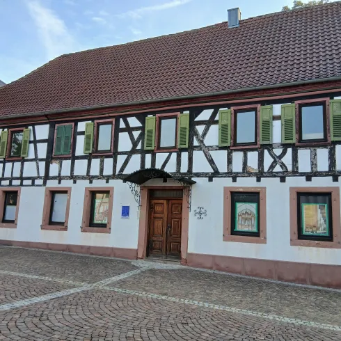 Ein traditionelles Fachwerkhaus mit grünem Fensterladen. Die Fassade ist weiß mit einem Ziegeldach und liegt an einem gepflasterten Platz.