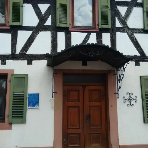 Ein traditionelles Fachwerkhaus mit weißen Wänden und grünen Fensterläden. Die Eingangstür aus Holz ist gut sichtbar unter einem Überstand.