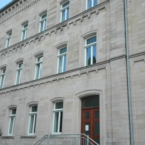 Ein historisches Gebäude mit einer hellen Sandsteinfassade und mehreren Fenstern. Der Eingangsbereich ist durch eine Treppe zugänglich.