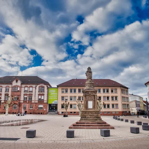 Ein schöner Platz mit einem Brunnen und einer Statue in der Mitte. Umgeben von historischen Gebäuden und einem beeindruckenden Himmel.