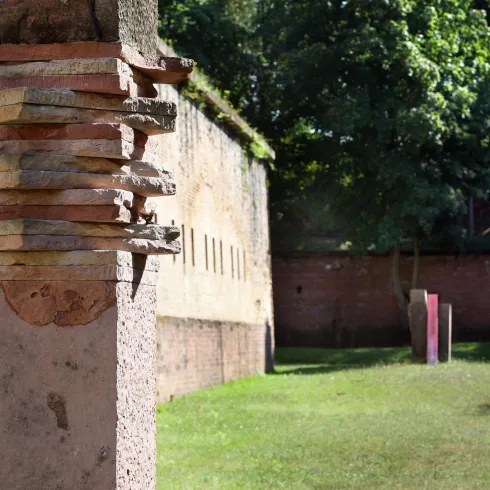 Ein historisches Gebäude mit einer Mauer und einem grünen Garten. Im Vordergrund steht ein verziertes Pfeilerstück.