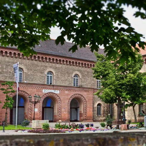 Ein historisches Gebäude mit einer schönen Fassade und großen Fenstern. Vor dem Gebäude befinden sich Blumenbeete und Bäume.