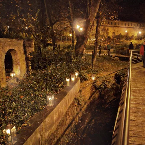 Eine romantische Nachtansicht mit einem beleuchteten Weg und Bäumen. Menschen gehen entlang der Holzbrücke, umgeben von kleinen Lichtern.